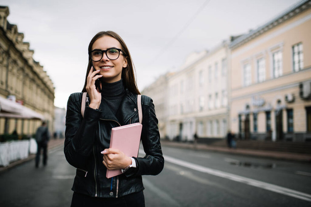 Millennial female student with education textbook using roaming internet connection for phone talking, happy tourist in optical eyewear making international conversation via smartphone application - Photo, Image