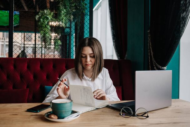 une femme d'affaires brune en chemisier blanc est assise à une table dans un café avec un ordinateur portable et des papiers et prend des notes - Photo, image