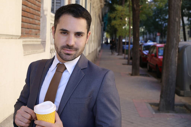 portrait of handsome young man in suit with tie holding paper cup of coffee on city street - Fotó, kép