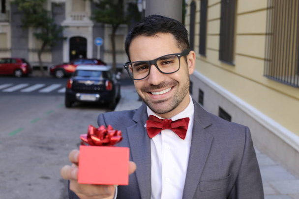Porträt eines hübschen jungen Mannes im Anzug mit roter Fliege, der eine rote Geschenkbox auf der Straße hält - Foto, Bild