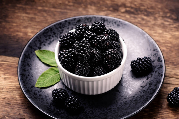 Fresh blackberries in a white bowl on a black plate on a wooden background close-up. - Photo, image