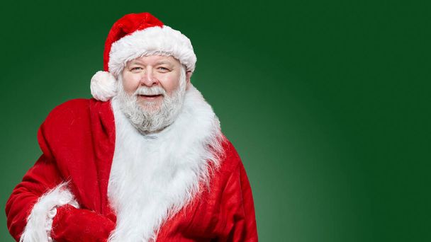 Nahaufnahme eines glücklichen Weihnachtsmannes in rotem Mantel und Hut, eine Tasche auf dem Rücken, isolierter grüner Hintergrund. - Foto, Bild