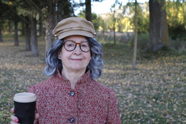 портрет пожилой женщины с седыми волосами, держащей бумажную чашку кофе на открытом воздухе - Фото, изображение