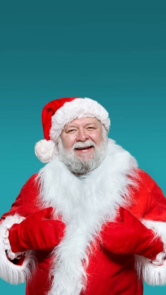 Zbliżenie uśmiechniętego Świętego Mikołaja w czerwonym płaszczu, rękawiczkach i kapeluszu, z podniesionymi rękami, odizolowane niebieskie tło. - Zdjęcie, obraz