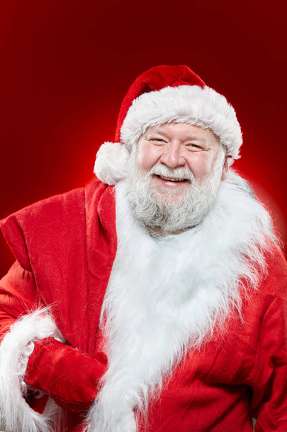 Счастливый Санта-Клаус одет в красное пальто и шляпу, держать мешок на спине, глядя на камеру, изолированный красный фон. - Фото, изображение