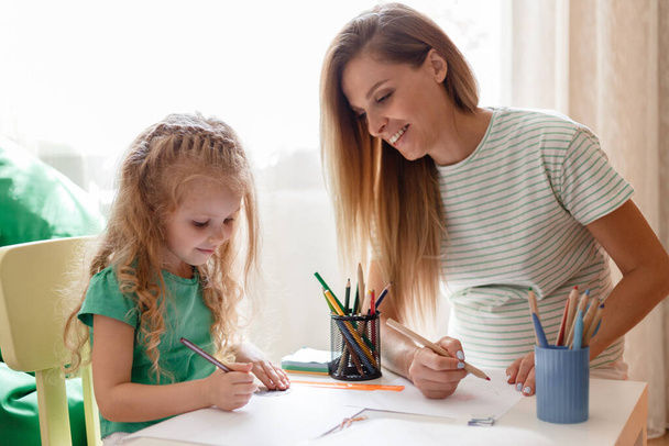 Portrait of mother and daughter drawing at home. Caucasian blonde pregnant mother helps to kid child preschool girl daughter doing homework, Spend Leisure time together play fun drawing.  - Foto, Imagen