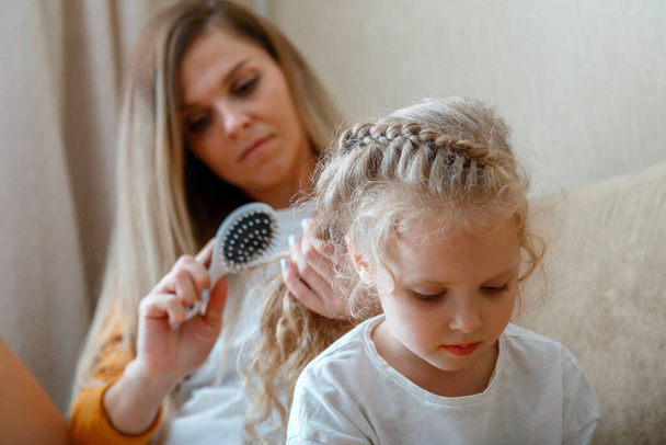 Mother combing daughter long hair by hairbrush. Caucasian blonde mom combing curly blond kid chaild. Mom does hair hairstyle and braids her braid. Womens morning routine.  - Foto, afbeelding