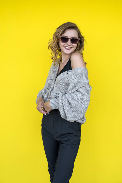 Modello femminile positivo in posa in maglia grigia maglione in occhiali da sole vicino alla parete gialla, all'aperto - Foto, immagini