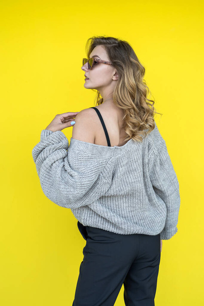Positive  female model  posing in gray knitted sweater in sunglasses near yellow wall, outdoor - Фото, изображение
