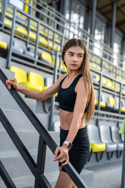 young slim female athlete having rest afret workout in sport clothing standing near colour sits at stadium  - Foto, Imagen