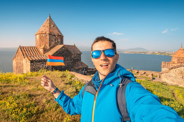De man met de Armeense vlag maakt selfie foto tegen de achtergrond van het Sevanavank klooster aan het Sevan meer. Vakantie en patriottisme concept - Foto, afbeelding