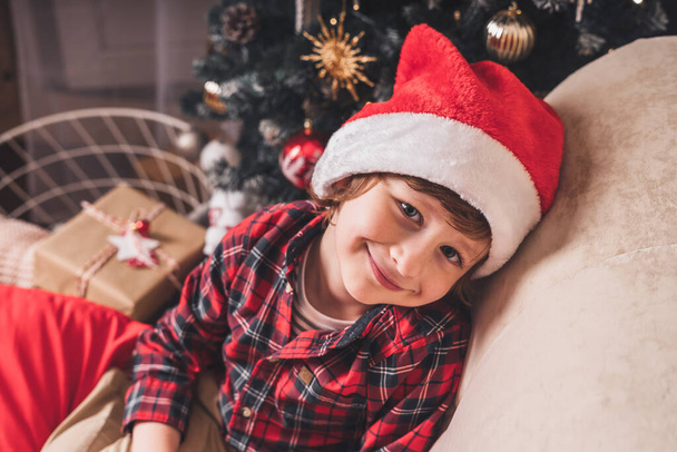 Χαριτωμένο μικρό χαμογελαστό παιδί αγόρι με κόκκινο καπέλο περιμένει τον Άγιο Βασίλη στο σαλόνι με χριστουγεννιάτικο δέντρο και δώρα στο σπίτι. - Φωτογραφία, εικόνα