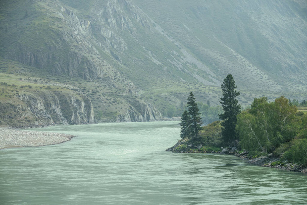 Прекрасний туманний гірський пейзаж з широкою гірською річкою. Темно-зелені похмурі пейзажі з великою гірською річкою в тумані. Темний вигляд на велику річку серед великих гір в дощову погоду
. - Фото, зображення