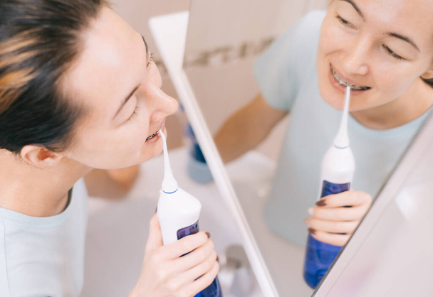 Woman with braces on her teeth brushing her teeth with by using a irrigate, before mirror, top view. - Foto, imagen