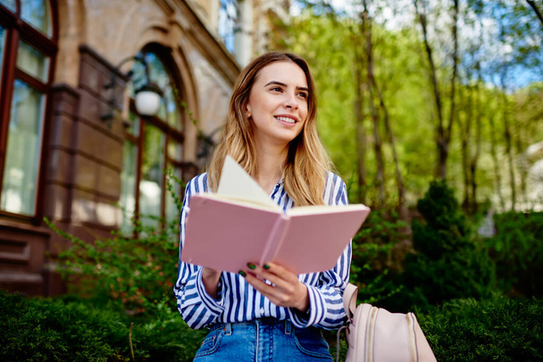 Happy youthful lady with blond hair wearing trendy clothes sitting with backpack against greenery in university yard and looking away while reading notes in planner - Foto, Imagen