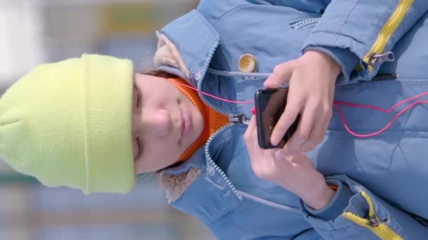 Вертикальное видео - девочка-подросток выбирает музыку в приложении для смартфона, вставляет наушник себе в ухо и наслаждается музыкой. - Кадры, видео