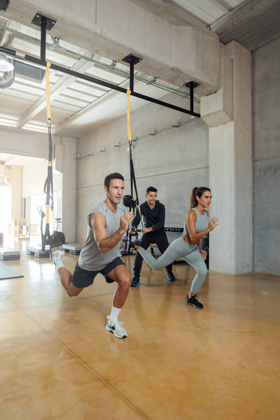 Suspension workout. Coach trains man and woman to do lunge exercise in a gym. - 写真・画像