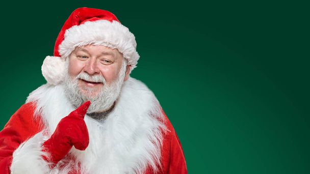 Kırmızı ceketli, eldivenli ve şapkalı mutlu bir Noel Baba 'nın ön yüzü. İşaret parmağıyla yeşil arka planı işaret ediyor. Metin için boşluk. - Fotoğraf, Görsel