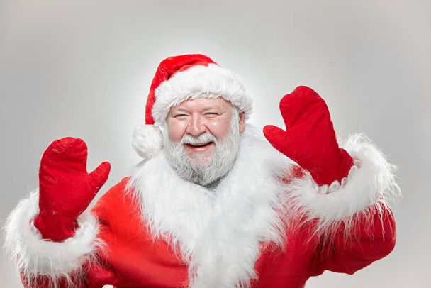 Перед нами зображення щасливого Санта Клауса, одягненого в червоне пальто з рукавицями і капелюхом, з піднятими руками високо ізольованим сірим фоном.. - Фото, зображення