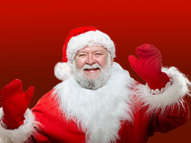 Close-up de uma imagem frontal de um Papai Noel feliz vestido com um casaco vermelho, com luvas e um chapéu, com as mãos levantadas alto fundo vermelho isolado. - Foto, Imagem