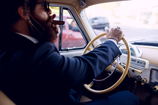 Close up de um homem barbudo, em óculos de sol e ternos elegantes pretos, posando cigarros fumantes sérios dentro do carro vintage, fundo ao ar livre. Gangster, conceito de máfia. Vista horizontal. - Foto, Imagem