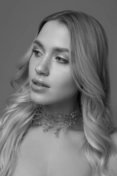 Portrait of a beautiful blonde with curly  long hair on a gray background with an ornament of small stones on her neck. Black and white photo. - Foto, imagen