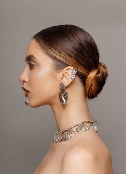 Portrait in profileof of a female model with splendid makeup, hairstyle, with naked shoulders, jewelry with ears, neck, nose, chin, over grey background. Vertical view. - Fotografie, Obrázek