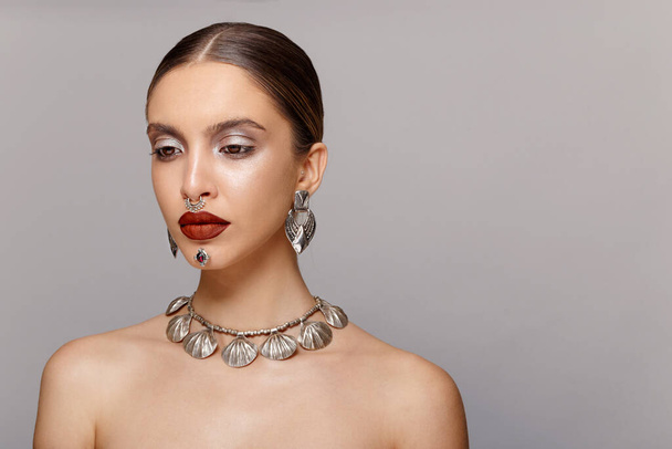 Beautiful with sensual and red lips, arranged, with jewelry, tight hair, looks down, with bare shoulders, over grey background. Horizontal view. Copy space. - Foto, afbeelding