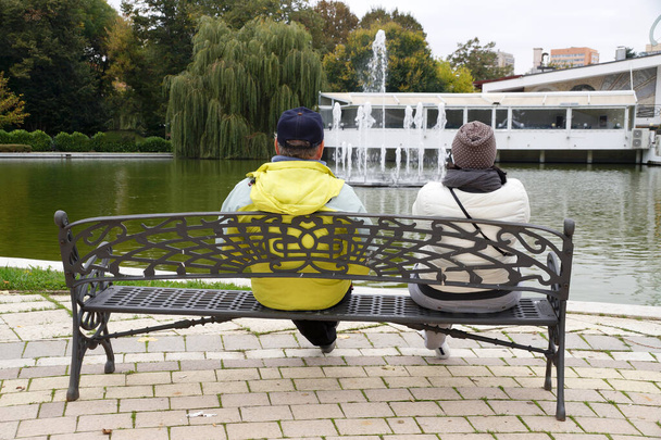 man and woman sit on a park bench and look at a pond with a fountain rear view - Valokuva, kuva