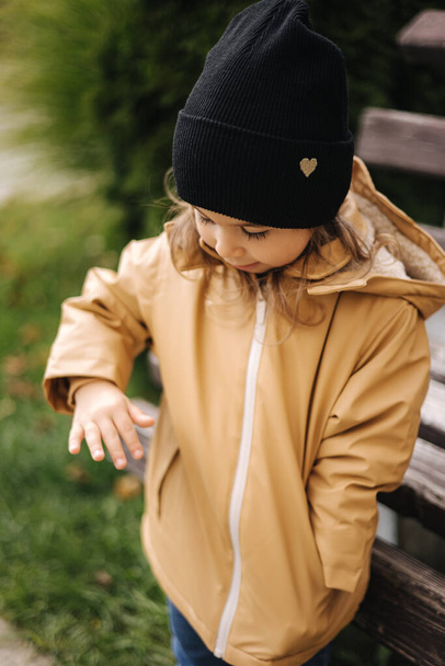 Feliz linda niña en chaqueta y sombrero posando al fotógrafo al aire libre por el banco. Pequeña modelo divertirse en otoño - Foto, Imagen