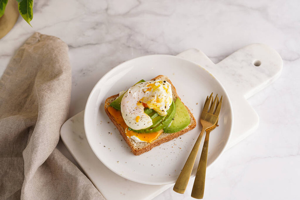 Colazione salata - Un pane tostato con crema di formaggio, avocado e uovo in camicia su un piatto bianco su vassoio di marmo - Foto, immagini
