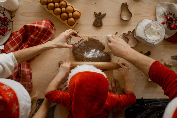 A top view of the hands of Mather, father and child in a santa hat, rolling dough with a rock on a wooden table. Flat lay. - Foto, afbeelding