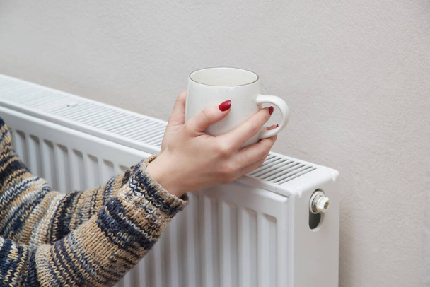Woman warming hands with tea cup and heating radiator.  - Photo, Image