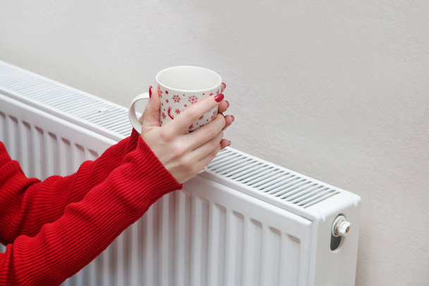 Nő melegítő kezét teáscsésze és fűtés radiátor.  - Fotó, kép