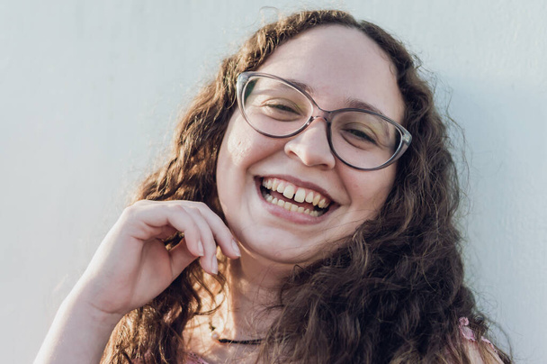 close up portrait of a pretty white hispanic latina girl with curls and glasses, smiling facing the camera, leaning against a wall, lifestyle concept and pride. - 写真・画像