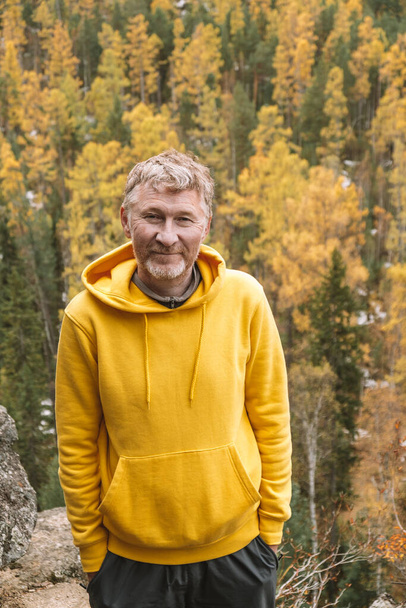 Un auténtico retrato de un viajero adulto sin afeitar sonriente en una sudadera con capucha amarilla en la cima de una montaña, sobre el fondo de un bosque otoñal. Enfoque selectivo. Primer plano. Medio ambiente sostenible. - Foto, Imagen