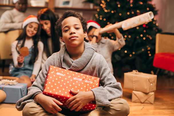 Szczęśliwy Afroamerykanin siedzi na podłodze w domu z prezentem świątecznym w rękach i patrzy w kamerę. W rozmytym tle jego rodzina otwiera prezenty. - Zdjęcie, obraz