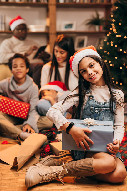 Egy boldog lány a Mikulás kalapjával a fején otthon ül egy karácsonyi ajándékkal a kezében. A homályos háttérben a multikulturális családja ajándékokat cserél.. - Fotó, kép