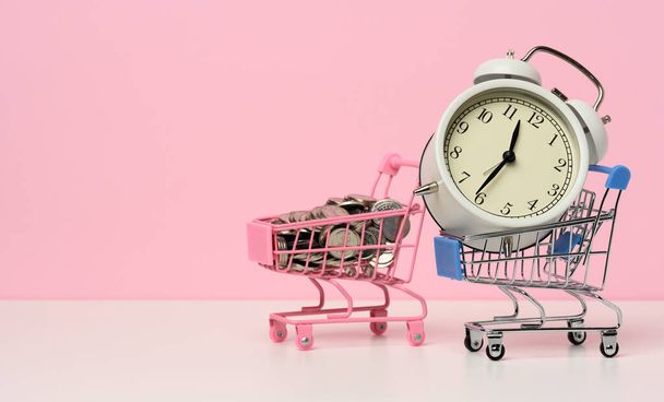 round alarm clock in a miniature shopping cart with change on a white table. Concept time is money, waste of money and poverty - Photo, Image