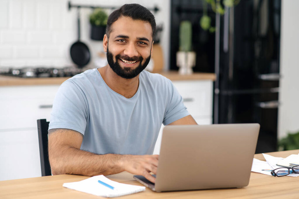 Retrato de belo indiano barbudo homem, freelancer, designer ou especialista em TI que trabalha em casa usando laptop, enquanto se senta à mesa na cozinha, olhando diretamente para a câmera e sorrindo amigável - Foto, Imagem