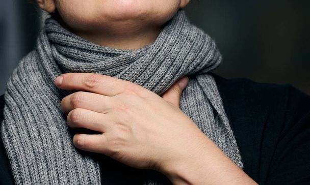 una bufanda gris se envuelve alrededor del cuello, y una mano sostiene la garganta. Frío y dolor de garganta - Foto, imagen