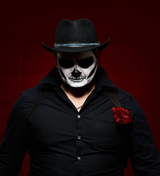  hombre está pintado con un esqueleto en un sombrero negro y camisa, cabeza abajo, fondo rojo - Foto, Imagen