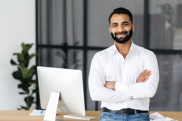 Porträt eines erfolgreichen intelligenten indischen Geschäftsmannes, Managers oder IT-Spezialisten, der ein weißes Hemd trägt, mit verschränkten Armen neben seinem Schreibtisch im Büro steht und freundlich in die Kamera blickt - Foto, Bild