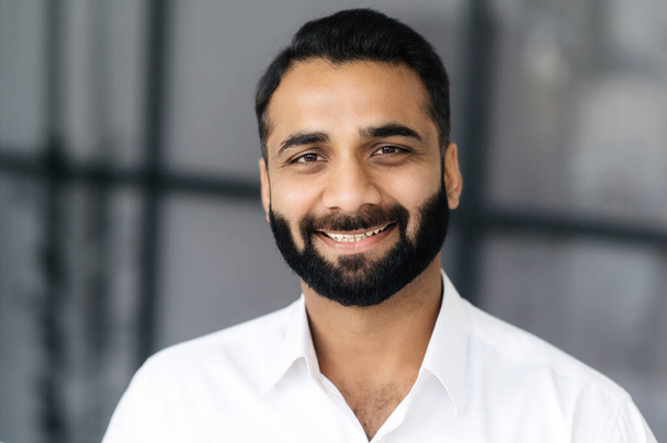 Retrato de close-up de um empresário indiano de barba inteligente de sucesso, gerente ou especialista em TI, vestindo uma camisa branca, olhando para a câmera com um sorriso amigável - Foto, Imagem