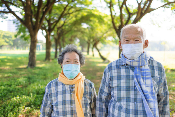 Ασιατικό ζευγάρι ηλικιωμένων με ιατρική μάσκα και φθινοπωρινό φόρεμα περπατώντας στο πάρκο - Φωτογραφία, εικόνα