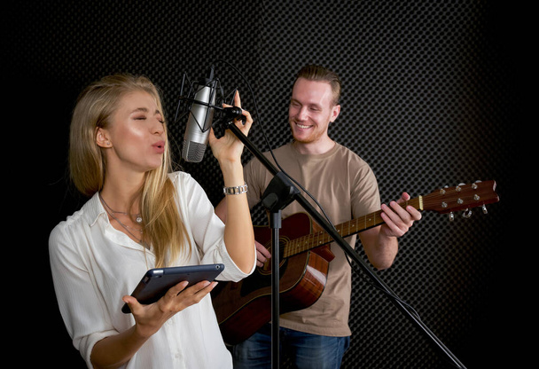 Jovem fêmea cantando com tablet na mão. Enquanto o homem tocando uma guitarra acústica na frente de paredes pretas à prova de som. Músicos que produzem música em estúdio de gravação profissional. - Foto, Imagem