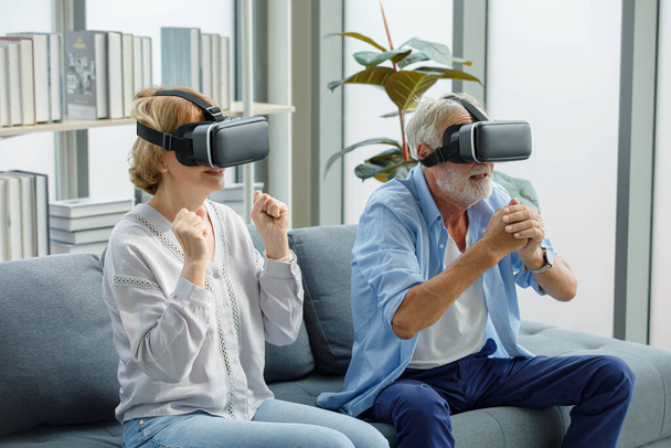 Un vieux couple de grands-parents âgés caucasiens porte un casque de réalité virtuelle. gris barbu et cheveux grand-père mari et grand-mère femme assis ensemble jouer vr jeu vidéo dans le salon. - Photo, image