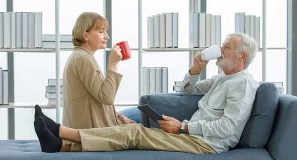 Caucasian old senior elderly happy lovely marriage couple grandparent gray bearded husband and pretty wife sitting relaxing resting on sofa smiling drinking cheers hot coffee cups together in morning. - Fotografie, Obrázek