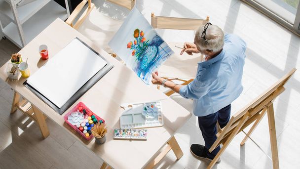Top view shot of Caucasian old senior elderly retired male painter spending time standing drawing painting beautiful flower bouquet in vase with colorful watercolor on paper alone in hobby workshop. - 写真・画像