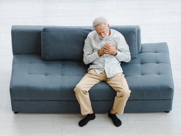 Кавказский пожилой пожилой седой волос и бородатый мужчина, лежащий на диване в гостиной один, держась за руки на груди, страдая от внезапной проблемы, болезненного сердечного приступа. - Фото, изображение